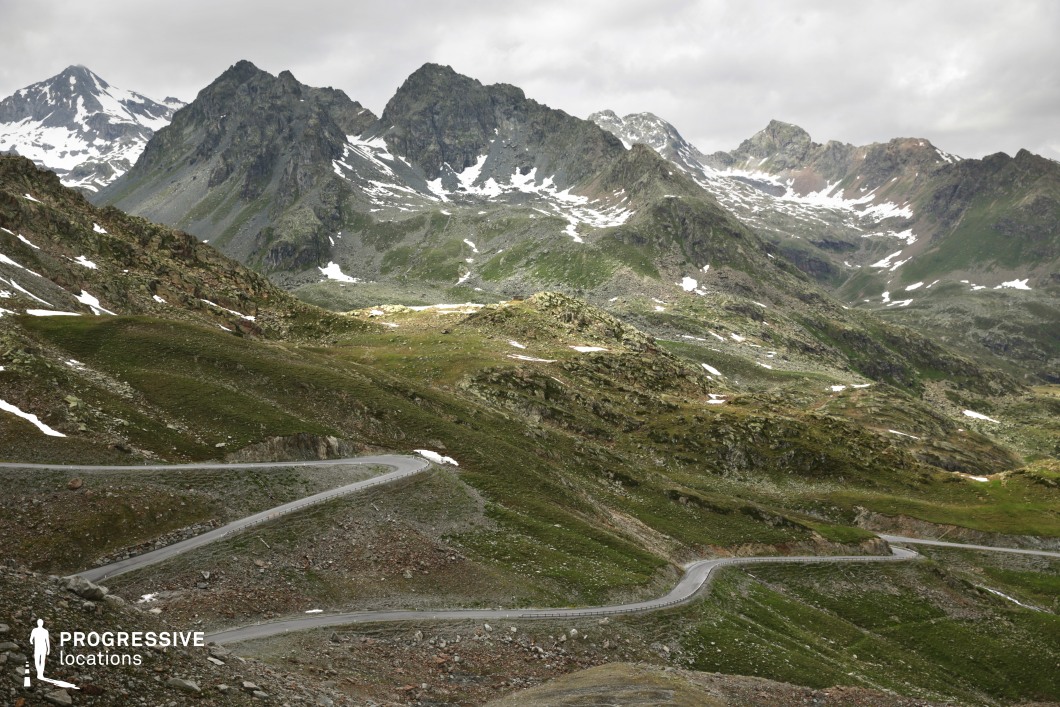 This remote mountainous location features a narrow serpentine road that weaves through rugged terrain, framed by steep, jagged peaks in the background. The landscape is composed of rocky outcrops, sparse patches of grass, and scattered snow deposits that emphasize the high-altitude character of the setting. The terrain exhibits strong geometric contours, with switchbacks creating dynamic diagonal lines that draw the eye across the frame. The visual composition benefits from a high vantage point, offering expansive depth and a layered backdrop of textured mountain faces. This location is ideal for filming adventurous or dramatic sequences, including car chases, exploration scenes, or scenic travel montages in genres such as thrillers, dramas, or documentaries, due to its raw, natural grandeur and challenging topography.