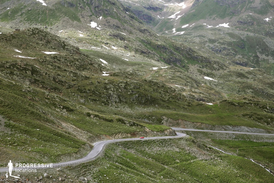 This expansive mountain location features a narrow, winding serpentine road that carves through a rugged alpine landscape. The terrain is composed of coarse, rocky slopes interspersed with patches of grass and sparse vegetation, lending the scene a raw, untamed character. The grey road contrasts sharply with the green and earthy tones of the mountainside, while its curvature and railing provide dynamic visual rhythm across the frame. The lack of dense structures or vertical elements emphasizes the vastness and isolation of the high-altitude environment. This location is especially suitable for filming sequences requiring dramatic natural backdrops, vehicular motion, or remote atmospheres, making it ideal for adventure, action, or introspective drama genres.