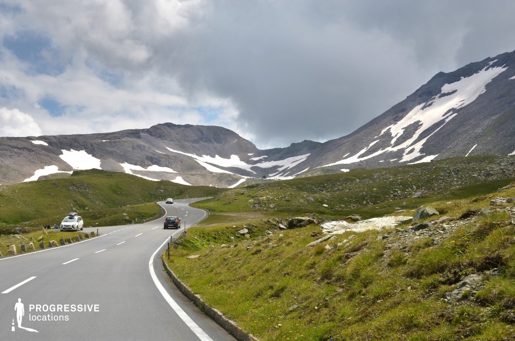 This high-altitude mountain plateau features a smoothly paved road winding through a vast, open alpine landscape, flanked by scattered rocks and grassy terrain. The backdrop is defined by broad, barren slopes with irregular snow patches clinging to the ridges and crevices, adding stark contrast to the muted grey rock formations. The landscape's color palette blends subdued greens with cool stone tones and white highlights, creating a visually raw and expansive atmosphere. The slightly elevated camera position captures the gentle undulations of the terrain and the road's curvature, enhancing depth and movement within the frame. The openness and minimalistic natural elements make this location particularly suitable for filming expansive, isolated sequences in road movies, psychological dramas, or commercials that require a sense of elevation, remoteness, or transition across rugged highland environments.