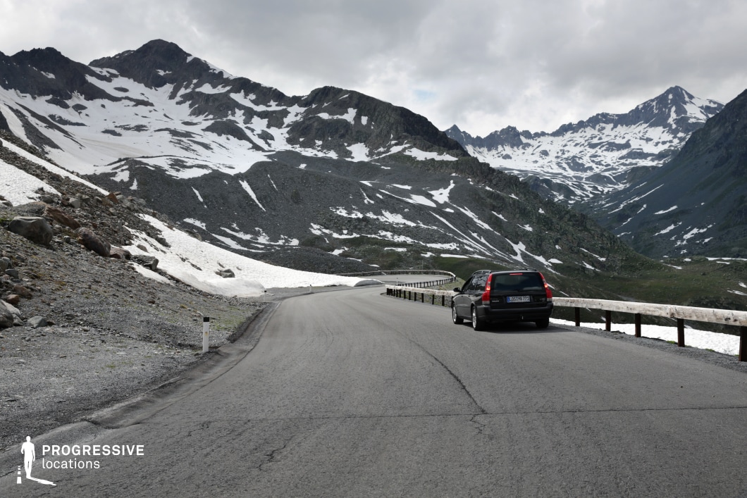This high-altitude mountain pass features a winding asphalt road flanked by a wooden guardrail and dramatic alpine terrain. The landscape is dominated by rugged slopes with a mix of exposed rock and lingering snow patches, creating a stark visual contrast of gray and white textures. The surrounding peaks rise sharply in the distance, forming a closed, bowl-like topography that enhances the sense of remoteness and scale. The road surface is wide and smooth, following a subtle curve that draws the eye into the mountainous horizon. The viewpoint is slightly elevated, with a perspective that accentuates the road’s integration into the harsh natural setting. This location offers a cinematic environment ideal for adventure, survival, or contemplative drama genres, with potential for dynamic vehicle shots, sweeping landscape visuals, and scenes emphasizing isolation or endurance in extreme conditions.