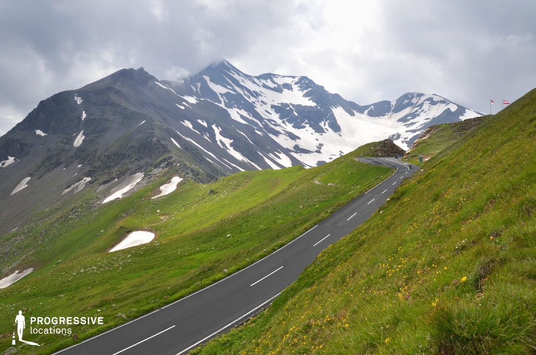 This striking location showcases a well-maintained, winding mountain road ascending through an expansive alpine landscape, flanked by steep green slopes dotted with wildflowers and remnants of snow. The perspective emphasizes the road’s curvature and gradient as it leads the viewer’s eye toward the towering, rugged mountain range in the background, partially blanketed in snow. The stark contrast between the vivid green foreground and the rocky, snow-striped peaks enhances the visual impact and highlights the location's dramatic elevation. The broad, open space and uninterrupted view of the mountains contribute to a sense of scale and remoteness. This location is ideal for filming high-altitude sequences, road journeys, or scenes requiring a vast and cinematic natural setting, especially for genres like adventure, drama, or documentaries.