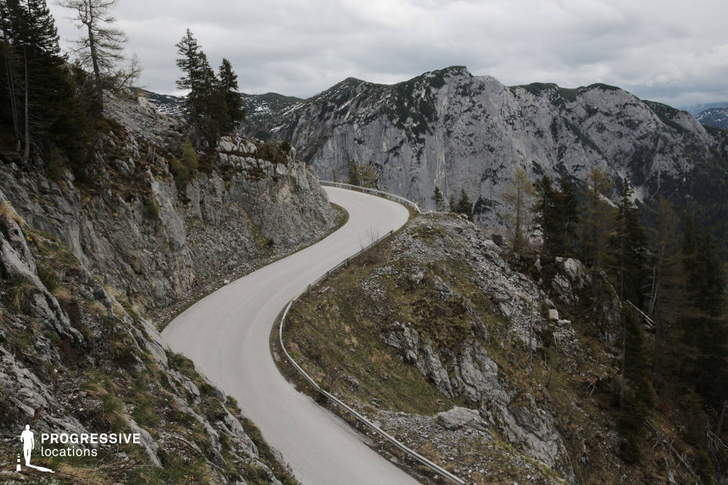 This striking location features a narrow, winding mountain road carved directly into rugged, rocky terrain. The light-colored asphalt contrasts sharply with the surrounding grey stone, emphasizing the tight, serpentine curves as the road ascends. On one side, steep cliffs rise dramatically, while the other drops into a forested valley with sharp elevation changes. Sparse alpine trees and craggy outcroppings reinforce the stark, untamed character of the environment. The backdrop includes massive vertical rock faces, creating a powerful sense of scale and seclusion. The elevated camera angle reveals both the geometric elegance of the road’s curve and the vertical drama of the landscape. This location is ideal for visually dynamic driving sequences, high-stakes chase scenes, or introspective travel moments in thrillers, dramas, and adventure films, offering a compelling mix of isolation, tension, and natural grandeur.