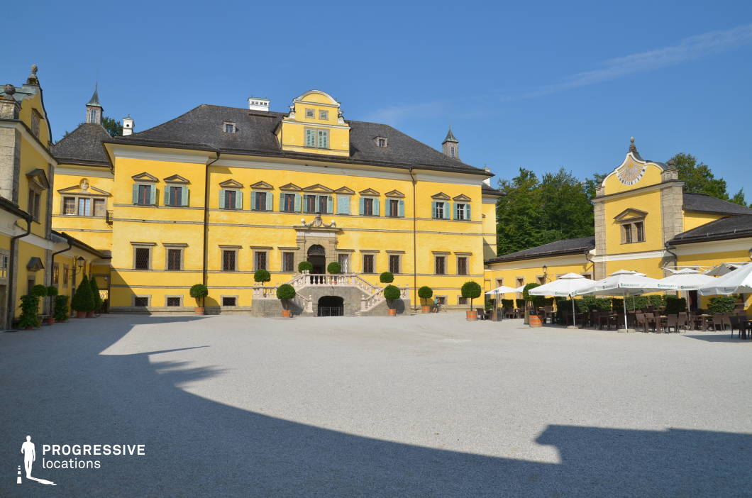 This expansive courtyard is enclosed by a vivid yellow Baroque-style palace, featuring symmetrical architecture, rustic window shutters, and a central staircase with ornate stone balustrades. The building’s façade is punctuated by repetitive geometric window frames and decorative stucco elements, enhancing the rhythmic visual structure. The open gravel-covered courtyard contrasts with the richly textured architecture, providing a clean, uncluttered foreground that emphasizes the stately presence of the building. On the right, a formal outdoor seating area with white parasols adds a functional layer, while potted topiary trees introduce controlled greenery. This location is well-suited for historical dramas, regal-themed productions, or elegant commercial shoots, offering a visually unified and architecturally striking environment for period or formal scenes.