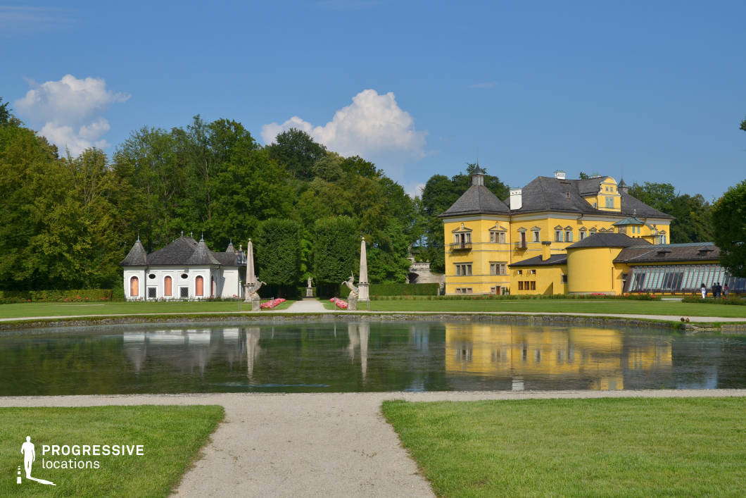 This visually striking location features a vivid yellow baroque-style palace framed by manicured gardens and a symmetrical reflecting pond in the foreground. The architectural composition is defined by the palace’s rounded tower elements, slate-tiled roofing, and rhythmic window placement, contrasted by a smaller, white neoclassical structure with a steep roof to the left. Two obelisks with sculptural bases serve as a visual gateway between the pond and the central garden path, guiding the eye toward the lush greenery and structured hedges beyond. The reflective water surface adds symmetry and depth to the scene, while the clear geometric layout enhances the formal elegance of the setting. This location is ideal for historical dramas, costume films, or luxury-themed productions, and it works especially well for establishing shots, romantic garden scenes, or refined cultural sequences due to its grandeur, balance, and stylistic richness.