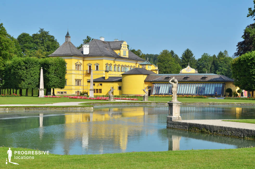 This scene presents a harmonious blend of classical architecture and landscaped garden design, centered around a large, reflective water basin bordered by manicured lawns and symmetrical gravel paths. The vivid yellow façade of the main building, with its multi-angled rooflines and turret-like projections, creates a bold visual contrast against the surrounding greenery. Marble statues positioned along the water’s edge add a sculptural rhythm to the composition, enhancing the formal garden aesthetic. The modern glass-panelled annex to the right introduces an architectural juxtaposition that expands the functional versatility of the site. This location is ideal for productions seeking an elegant, historical European estate atmosphere, suitable for romantic, period, or cultural genre films, as well as promotional visuals requiring serene, balanced compositions with architectural and sculptural sophistication.