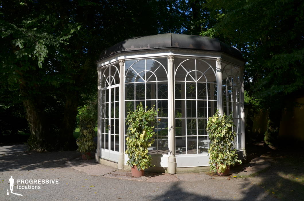 This location features a small, elegant glass pavilion set within a shaded park environment. The structure is octagonal with tall, white-framed arched windows and classical decorative columns that add a refined, almost romantic architectural tone. Its metal-framed, slightly domed roof shows signs of patina, enhancing its historical character. Surrounded by mature trees and a gravel path, the setting is tranquil and secluded, offering a mix of natural shadow and soft, filtered light. The pavilion's symmetry and transparency allow for both interior and exterior shots with visual continuity. Ideal for period dramas, romantic or introspective scenes, and fashion shoots, the location offers a versatile backdrop with a timeless aesthetic.