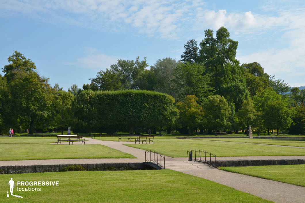 This expansive formal park is defined by its geometric layout of trimmed grass fields, intersecting gravel pathways, and evenly spaced wooden benches, offering a clear and symmetrical spatial composition. A narrow stone-lined canal runs through the middle of the scene, crossed by a small decorative footbridge with simple metal railings that adds structural rhythm. The background is framed by a dense mix of mature trees, including meticulously shaped hedge-like forms and taller, natural-growth varieties, creating a strong contrast between order and organic elements. The overall setting feels open yet structured, making it highly adaptable for filming tranquil, contemplative, or ceremonial scenes. The unobstructed sightlines and consistent greenery provide a versatile location for dramas, art films, or commercial productions seeking an elegant, serene outdoor setting.
