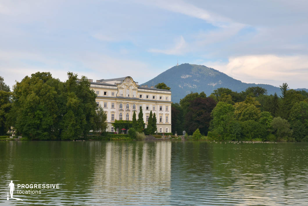 This image captures a grand Baroque-style palace situated directly on the edge of a tranquil lake, framed by lush, mature trees and backed by a prominent forested mountain. The building’s light-colored façade features symmetrical windows, ornate detailing, and a central gable with classical ornamentation, contributing to an overall refined and historic atmosphere. The reflective lake surface introduces a strong visual axis, emphasizing the symmetry and elegance of the architecture. The scene is dominated by natural tones—greens of varying saturation, soft beiges, and the deep blues of the mountain—creating a serene and cinematic palette. The palace appears to be four stories tall and is visually anchored by tall cypress-like trees and structured hedges. This location offers a highly versatile backdrop for historical, romantic, or period film genres, as well as scenes requiring a stately, secluded, or aristocratic ambiance. Its architectural richness and natural framing make it especially suitable for establishing shots, tranquil sequences, or introspective moments.