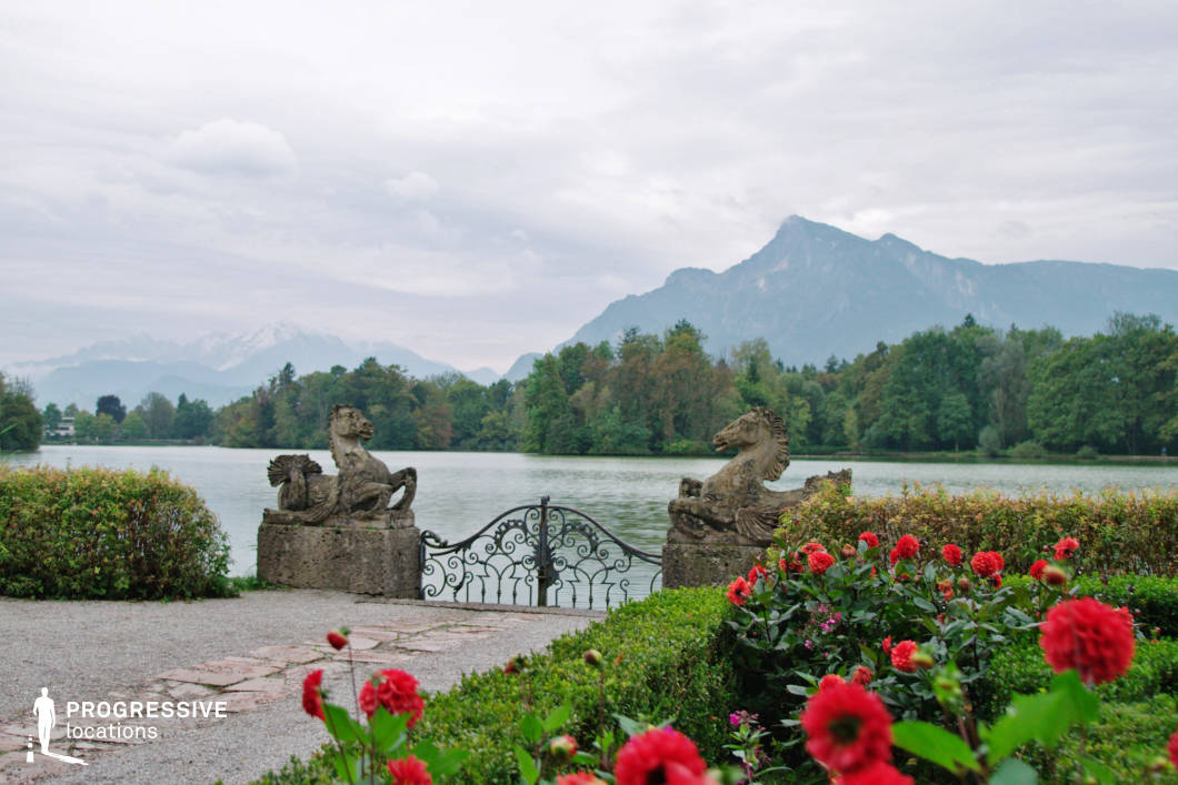 This elegant lakeside terrace opens to a symmetrical wrought-iron gate flanked by two large stone horse sculptures, offering a commanding focal point at the water's edge. The foreground is lined with well-maintained box hedges and vivid red flowers, leading to a gravel path and natural stone pavers that frame the approach. The calm lake stretches across the middle plane, bordered by a dense, mature tree line that introduces deep greens and earthy tones, while the background reveals a dramatic mountain range with layered peaks that enhance the sense of scale and depth. The sculptural elements, ornamental ironwork, and manicured landscaping evoke a refined, almost aristocratic ambiance. Ideal for period dramas, historical narratives, romantic scenes, or visually rich establishing shots, this location’s blend of classical design and natural grandeur offers versatility and cinematic visual impact.