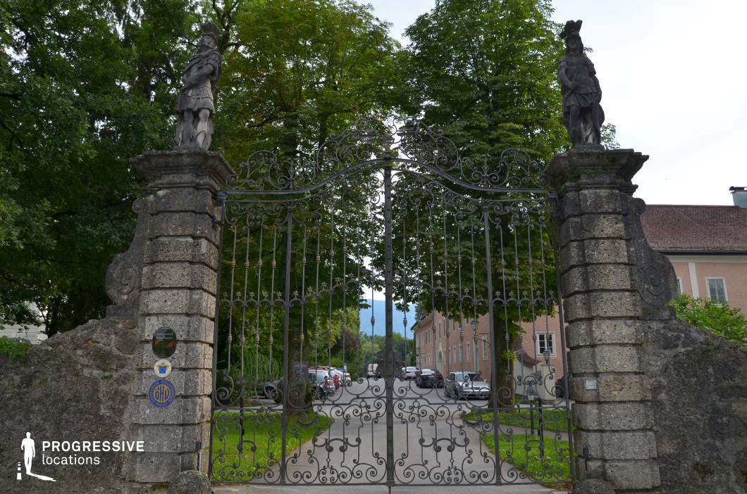 This visually striking entrance features a pair of tall, weathered stone pillars supporting an elaborate wrought iron gate adorned with swirling patterns and classical detailing. Atop each pillar stands a sculpted stone figure in historic military attire, adding a monumental and authoritative presence to the composition. The gate opens onto a tree-lined avenue flanked by pastel-toned classical buildings with rhythmic window arrangements and gabled roofs. The scene is framed symmetrically from a frontal, eye-level camera angle, emphasizing balance and depth. This location offers a strong visual identity and historical ambiance, making it suitable for period dramas, fantasy, or aristocratic settings, particularly for establishing shots, ceremonial entrances, or moments requiring grand architectural framing.