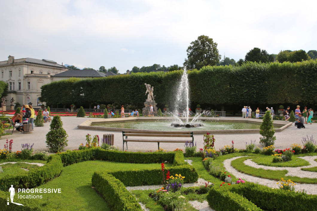 This image showcases a meticulously designed Baroque-style garden featuring symmetrical gravel paths, ornate hedgerows, and vibrantly planted flowerbeds, all leading toward a centrally placed circular fountain with a vertical water jet. The garden is enclosed by a dense, carefully trimmed hedge wall that creates a strong sense of geometric enclosure and symmetry. A stone statue atop a sculptural pedestal adds vertical interest and classical character to the space, while the corner of an elegant historic building in light stone with neoclassical elements anchors the background. The garden’s dominant palette consists of greens, beiges, and floral accents, fostering a balanced and refined aesthetic. This highly structured environment, with its ornamental layout and architectural context, offers an ideal backdrop for period dramas, historical reenactments, or stylized romantic scenes, especially where elegance, grandeur, and symmetry are essential visual themes.