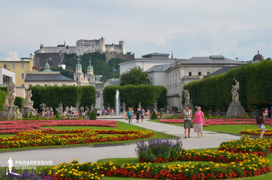 This location features a meticulously maintained baroque-style formal garden framed by symmetrical gravel pathways, vibrantly patterned flowerbeds, and a series of expressive stone sculptures on pedestals. Lush, geometrically trimmed hedges and mature trees border the central walkway, creating strong visual depth that guides the eye toward a prominent neoclassical fountain at the composition’s midpoint. In the distance, the scene is anchored by an imposing hilltop fortress and the twin-domed towers of historic ecclesiastical architecture, offering a powerful contrast between structured greenery and fortified medieval stonework. The visual layering of garden, urban architecture, and elevated fortress creates a rich, three-dimensional backdrop. This location is highly suitable for period films, costume dramas, historical documentaries, or high-end fashion shoots requiring a regal, cultured ambiance with classical European elegance.
