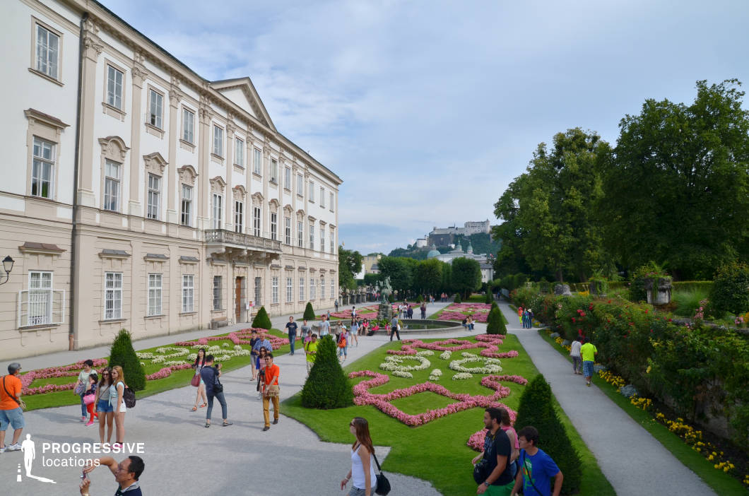 This location features a symmetrical baroque garden adjacent to a grand neoclassical palace, characterized by its cream-colored façade, decorative window pediments, and orderly architectural rhythm. The garden showcases meticulously arranged geometric flowerbeds bordered by manicured pathways, with sculpted bushes accentuating the formal layout. The area is enclosed by tall, mature trees and dense hedgerows, adding vertical contrast and visual depth to the otherwise horizontal composition. A distant fortress rises above the background, enhancing the historical and European urban character of the setting. The camera angle captures a wide perspective from an elevated position, emphasizing the axial alignment of the garden and the visual connection between architecture and landscape. This location is ideal for filming scenes requiring a refined, elegant, and structured outdoor setting—well-suited for genres such as historical drama, romance, or period pieces, as well as formal event or ceremonial scenes.