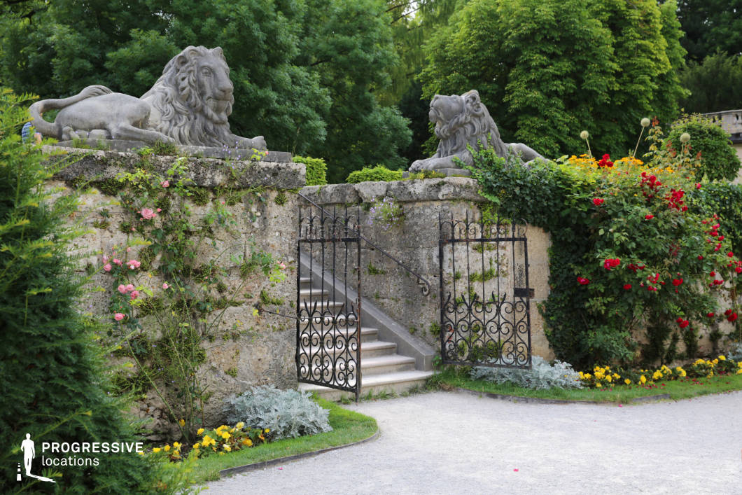 This location features a historical garden entrance framed by weathered stone walls and crowned with two imposing stone lion sculptures. A short staircase with wrought iron railings leads through an open, decorative iron gate, inviting movement into the upper terrace. The stonework displays aged textures with moss and climbing plants, enhancing the timeless, slightly romantic atmosphere. Surrounding flowerbeds add vivid color contrast with blooming red, yellow, and pink flowers, while dense greenery in the background provides a natural enclosure. The symmetrical layout, sculptural elements, and formal garden details create a refined and classical ambiance, ideal for period dramas, historical settings, or elegant garden scenes in film and photography.