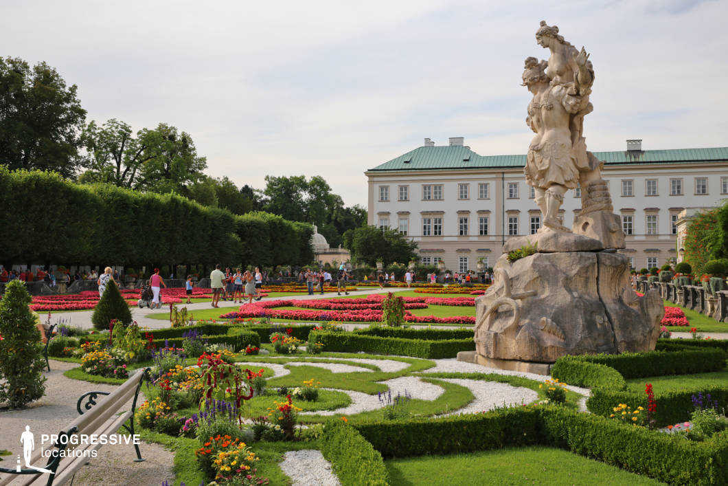 This meticulously landscaped baroque garden features a symmetrical design with vibrant, color-coordinated flower beds bordered by well-maintained low hedges and winding gravel paths. Dominating the foreground is a large, ornamental stone statue atop a rugged pedestal, which serves as the central sculptural element. The formal layout is complemented by a white neoclassical building in the background, characterized by its pale facade, regular window pattern, and muted green roof, contributing to an elegant, aristocratic atmosphere. Geometric planting patterns and precisely trimmed trees enhance the visual structure, offering depth and balance across the frame. The space’s organized, refined aesthetic, along with its clear sightlines and historical ambiance, makes it ideal for filming romantic scenes, period dramas, or upscale promotional content requiring classical or European garden settings.