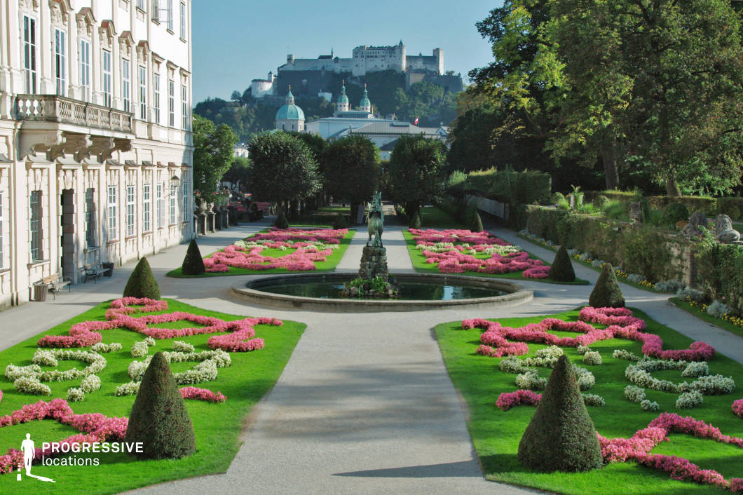 This image showcases a meticulously maintained formal garden with symmetrical design, dominated by vibrant pink and white flowerbeds arranged in ornamental patterns on a backdrop of evenly cut green lawns. The central pathway is flanked by trimmed conical shrubs and leads to a small circular fountain featuring a classical equestrian statue, serving as the focal point of the composition. To the left, a pale stone façade with tall sash windows and decorative balustrades enhances the historical elegance of the scene, while the structured layout and precisely pruned greenery underscore the garden’s Baroque character. In the distance, a fortress-topped hill adds vertical contrast and depth to the composition, enhancing the spatial layering and giving a strong sense of place. The camera angle emphasizes symmetry and depth, making this a prime location for period films, costume dramas, or romantic scenes requiring a sophisticated, timeless European setting.