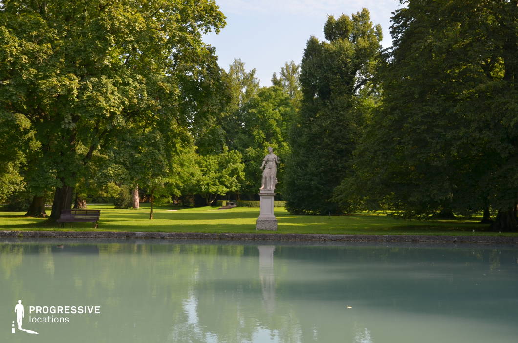 This location features a manicured park setting dominated by dense, mature trees and a calm, pale-toned pond in the foreground. The centerpiece is a classical stone statue on a pedestal, symmetrically framed by the surrounding greenery. The park’s lawn is neatly maintained, offering a flat, uninterrupted expanse that contrasts with the dynamic verticality of the tree canopy. The materials range from carved stone to natural elements like grass and water, creating a serene yet structured atmosphere. The geometric symmetry of the composition, with the statue as the focal point and its reflection on the pond’s surface, adds a refined visual depth. This location is ideal for filming scenes that require a timeless, elegant, or contemplative ambiance, and it is particularly suited for historical, romantic, or fantasy genres.
