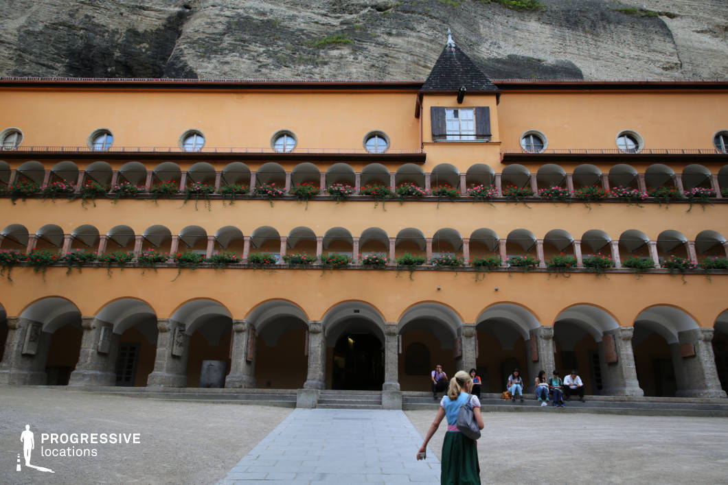 This striking location features a symmetrical courtyard facade with three levels of uniform archways and colonnades, all painted in a warm ochre hue. Each balcony level is decorated with flower boxes, adding vibrant accents against the stone and plaster textures. The ground level rests on robust stone pillars, forming a sheltered arcade that supports the rhythmic visual repetition above. A small tower-like projection with shuttered windows adds architectural variety near the center of the roofline. The entire structure is set directly against a steep natural rock wall, creating a dramatic vertical contrast between built and natural elements. This setting is visually cohesive, texturally rich, and architecturally unique—making it highly suitable for historical dramas, institutional settings, or scenes requiring cloistered, introspective atmospheres with a strong sense of place.