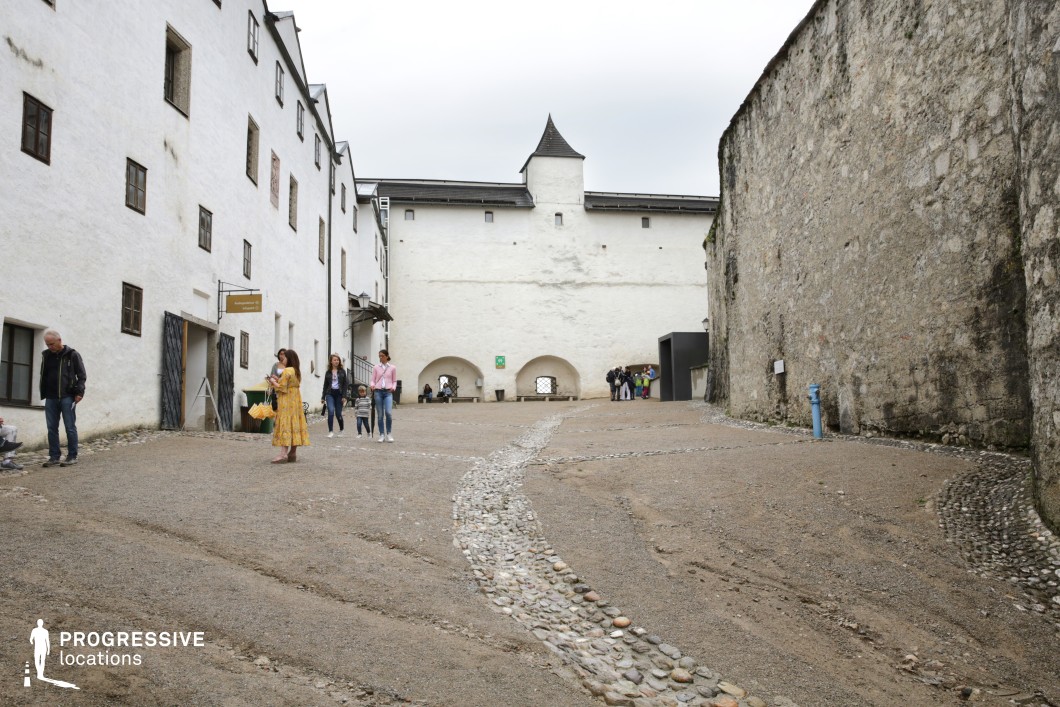 This enclosed courtyard is defined by tall, austere castle walls constructed from rough-hewn stone and whitewashed masonry, creating a strong visual contrast between the surfaces. The sloped ground is covered with gravel and segmented by winding bands of embedded river stones, guiding the viewer’s eye toward a fortress-style gate with three arched openings. The walls feature small, irregularly placed windows, enhancing the historical authenticity and fortified feel of the setting. The dominant architectural style is medieval, characterized by minimal ornamentation and practical design. The high vantage point of the camera emphasizes the spatial containment and verticality of the walls, contributing to a sense of seclusion and historical gravity. This location is highly suitable for period pieces, historical dramas, or productions requiring a fortress or castle environment with authentic textures and architectural depth.