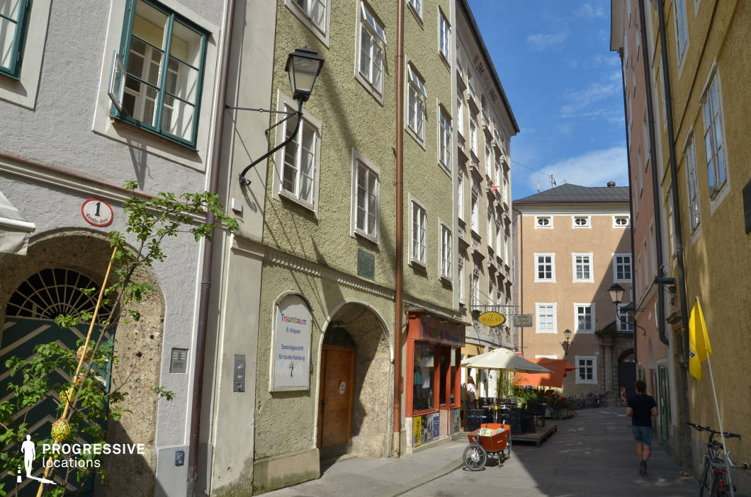 This intimate urban alley is enclosed by multi-story buildings with textured stucco facades in varied muted tones, including sage green, ochre, and cream, contributing to a cohesive yet lively palette. The narrow street features traditional architectural elements such as arched doorways, wrought-iron signs, and classic window shutters, while modern storefronts are discreetly integrated into the ground level. The perspective emphasizes verticality and linear depth, accentuated by the converging lines of the buildings and the unobstructed central path. Hanging lanterns and subtle decorative details lend a timeless European charm. The confined yet visually rich setting is ideal for filming character-driven scenes, dialogue sequences, or transitional shots in period films, romantic dramas, or travel-related content requiring an authentic old-town ambiance with a walkable, human-scale atmosphere.