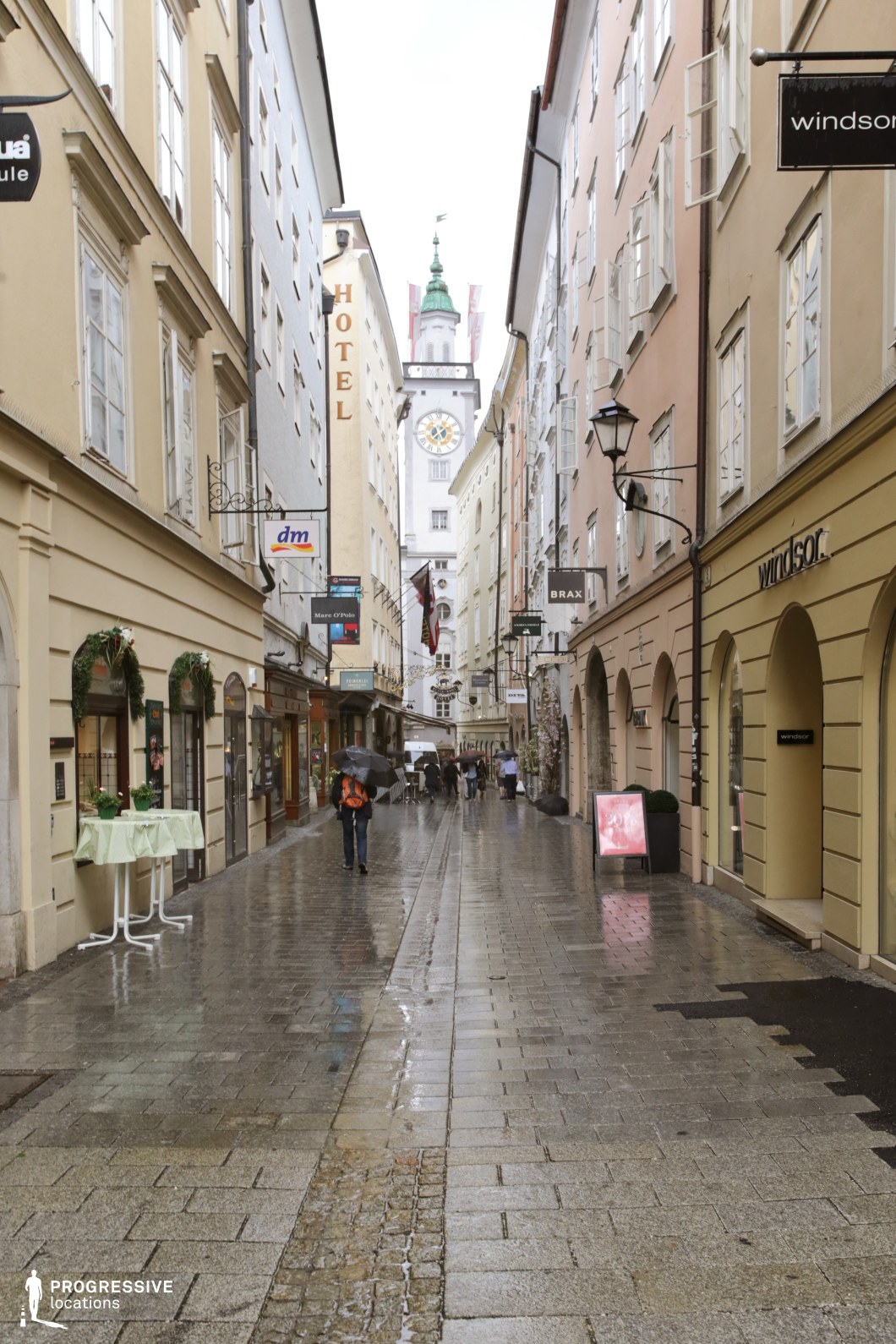 This image captures a narrow urban street framed by two contrasting building styles leading to a grand baroque church at the focal point. On the left, cream-colored façades adorned with ornate wrought-iron balconies exhibit a classic, historic European aesthetic. The right side features arched windows with iron bars on a simpler, yet elegant neoclassical structure. The central church, with its intricate yellow and white exterior, twin green domes, and clock towers, dominates the perspective, adding a dramatic and formal element. The cobblestone street, with its aligned bollards, creates a sense of order and guides the eye naturally toward the church. This location is ideal for scenes requiring a blend of historical charm and urban intimacy.