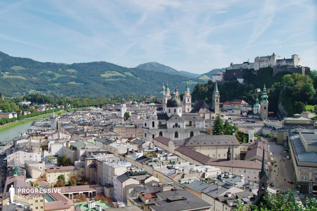 This panoramic urban vista reveals a densely built baroque city core nestled in a river valley, framed by forested hills and expansive mountain ridges. Dominating the skyline are domed churches and spired towers with green oxidized copper roofs, contrasted by the pale stone façades and symmetrical rooflines of the surrounding residential and civic buildings. Perched on a steep wooded outcrop in the background, a large fortress complex adds a sense of historic gravity and visual hierarchy to the scene. The visual depth is enhanced by the gentle curvature of the river to the left and the layering of rooftops, which leads the eye naturally through the frame. This elevated perspective captures both architectural richness and geographical context, making the location highly suitable for historical, cultural, or documentary productions, as well as for wide establishing shots in narrative films seeking a classical European urban atmosphere.