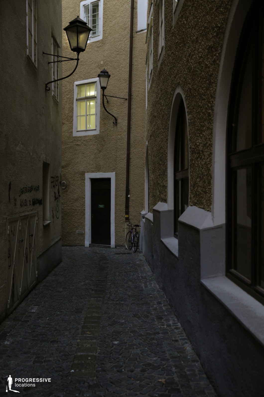 This secluded alleyway is framed by tall, textured façades in muted tones, with rough stucco and simple white-framed windows contributing to a restrained historic character. The uneven cobblestone paving, worn with age, adds depth and texture to the narrow passage, which curves gently and leads the eye toward a modest dark door at the end. Wall-mounted street lamps with wrought iron fixtures, along with arched windows on one side, accentuate the old-world atmosphere. The spatial proportions are tight and vertically enclosed, emphasizing a sense of intimacy and seclusion. This location is ideal for filming scenes requiring urban tension, mystery, or historical ambiance, making it especially suitable for thrillers, noir films, or character-driven dramas.