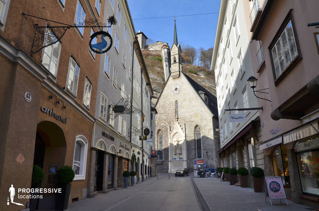 This urban location captures a narrow, stone-paved street flanked by tall, pastel-toned historic buildings that create a canyon-like architectural effect. The facades are textured with traditional stucco and regularly spaced, white-framed windows, while wrought-iron hanging signs add visual interest and an old-world character to the retail fronts. At the visual terminus stands a vertically emphasized stone church with a sharp spire and gothic-style entrance niche, integrated into the rising rock face behind it. The composition draws the viewer’s eye along the linear perspective of the street, concluding at the ecclesiastical structure which anchors the frame. This compact yet visually rich environment offers a well-preserved old-town ambiance, ideal for scenes requiring a European heritage setting, upscale commercial atmosphere, or visually contained street-level sequences in historical or cultural productions.
