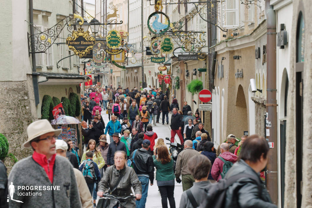 This image captures a narrow, elongated pedestrian street flanked by traditional multi-story façades with arched entrances, textured stonework, and neutral-colored plaster walls. The standout visual element is the dense array of intricate wrought-iron shop signs that project from the buildings in decorative patterns, featuring gold detailing, ornamental scrolls, and historic emblems. These signs form a continuous line overhead, adding rich vertical layering and evoking a distinctly old-world European charm. The tightly packed architecture and limited sky visibility create an enclosed, intimate atmosphere, while the linear composition draws attention along the depth of the street. Ideal for filming historical or culturally rich urban scenes, this location suits genres such as drama, romantic comedies, and period pieces, especially those seeking a classical European shopping district or atmospheric old-town setting.