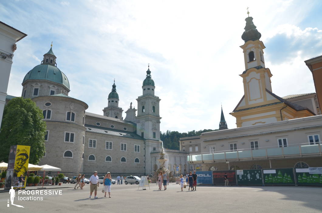 This expansive urban square is defined by grand Baroque architecture, with the dominant structure being a cathedral complex characterized by rounded towers, arched windows, and green copper domes and spires. The surrounding buildings present a mix of pale stone facades and ornate decorative elements, offering a cohesive historical aesthetic. The square is paved and open, enhancing the monumental scale of the structures, while the centralized fountain adds a vertical focal point and classical detail. Photographed from a slightly low, frontal perspective, the image emphasizes the verticality of the towers and the openness of the plaza. The location's strong architectural character and clean sightlines make it ideal for historical films, period dramas, and formal urban sequences where a sense of European grandeur and cultural context is desired.