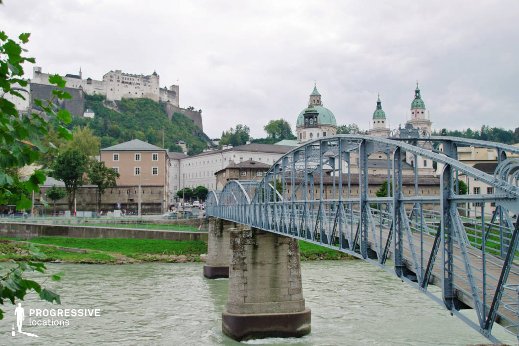 The image presents a dynamic urban composition anchored by a steel pedestrian bridge with arched trusses and robust stone piers crossing a wide, flowing river. This functional structure introduces a distinctly industrial contrast to the historic architecture behind it, including domed churches and baroque buildings characterized by light facades and green copper rooftops. The elevated fortress dominating the hilltop background adds a medieval layer to the multi-era skyline, contributing to the scene's strong historical narrative. The overall setting is geometrically rich, with linear bridge elements intersecting the vertical and curved forms of the older architecture, offering compelling depth and perspective. This location is ideal for dramatic or romantic urban scenes, architectural documentaries, or travel-themed productions due to its layered visual history, architectural variety, and strong spatial structure.