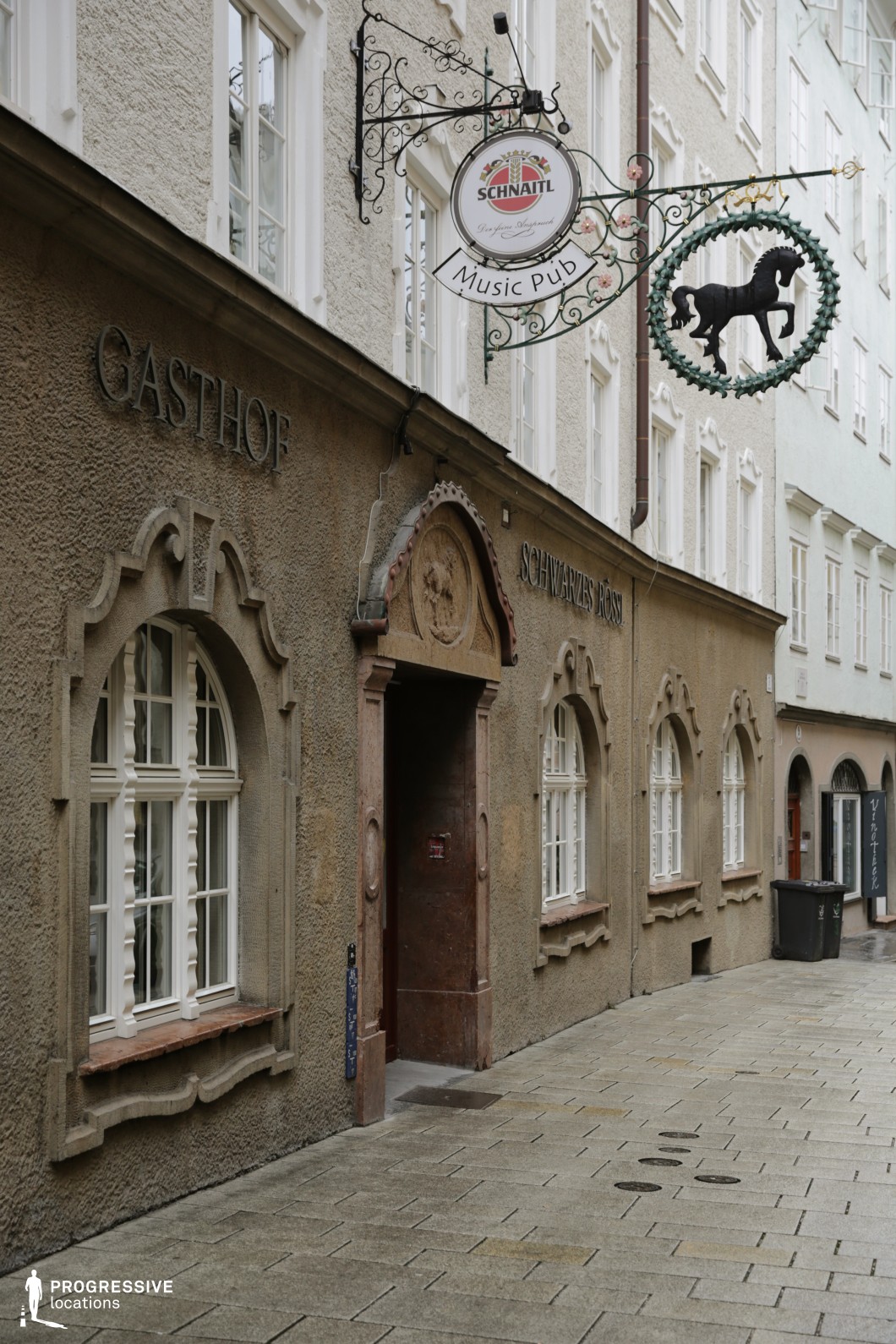 This intimate urban location features a narrow, stone-paved alley flanked by textured stucco buildings in muted earth tones. The main facade presents traditional architectural details, including arched windows with sculpted frames, a decorative doorway topped with a relief, and wrought iron signage extending outward, adding vertical interest. White-framed windows with old-style glazing bars align rhythmically along the wall, emphasizing the historical character of the structure. The tight passage and frontal, slightly angled camera perspective accentuate the narrowness and the verticality of the building features. Ideal for filming scenes that require old-world charm or European urban authenticity, this location suits genres such as historical drama, mystery, or romance, and works well for walk-and-talk sequences or atmospheric establishing shots.
