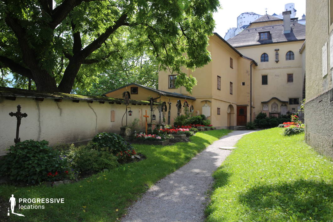 This intimate location features a quiet gravel path winding through a well-maintained monastery garden, bordered by manicured grass and colorful flowerbeds. A low plastered wall lines one side, adorned with wrought iron crosses and lanterns, contributing to a solemn and contemplative atmosphere. The buildings exhibit simple, historic architectural forms with ochre-toned facades, small wooden windows, and traditional pitched roofs. The presence of religious reliefs and inscriptions further reinforces the spiritual character of the space. Framed by large, mature trees and shielded from the outside world, this setting is ideal for contemplative, historical, or character-driven scenes in dramas or period films requiring a serene, enclosed environment.