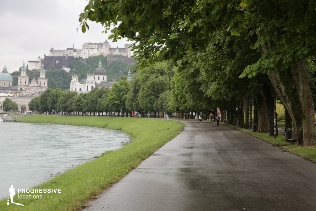 This long, gently curving riverside promenade is defined by its dual elements: a narrow green embankment bordering calm waters on one side and a dense, symmetrical line of mature trees on the other, creating a natural canopy over the paved path. In the background, a dramatic hillside is crowned by an expansive stone fortress, while below it, domed churches and Baroque façades establish a rich architectural skyline. The composition balances natural and historical elements, with strong linear geometry from the path and tree alignment enhancing visual depth. This location is particularly suitable for period dramas, romantic scenes, or atmospheric city walks, offering a blend of urban elegance and scenic tranquility.