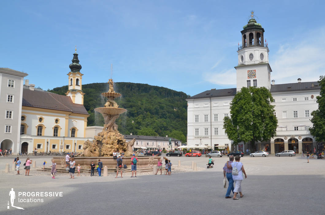 This image presents a spacious, open town square defined by historical architecture and dominated by an ornate multi-tiered Baroque fountain constructed from sculpted stone. The buildings surrounding the square exhibit a blend of neoclassical and traditional European elements, with pale facades, rhythmic window placements, and distinctive towers—one with a decorative belfry and clock. The square is paved with fine gravel, creating a uniform surface that enhances the clarity of the architectural lines and the central fountain’s sculptural detail. The visual composition benefits from the backdrop of a densely forested hillside, adding depth and contrast to the otherwise urban setting. This location is ideal for a wide range of film productions including historical epics, period dramas, and cultural documentaries, particularly those requiring an iconic European town square with strong architectural character and flexible spatial dynamics for crowd scenes or large-scale setups.