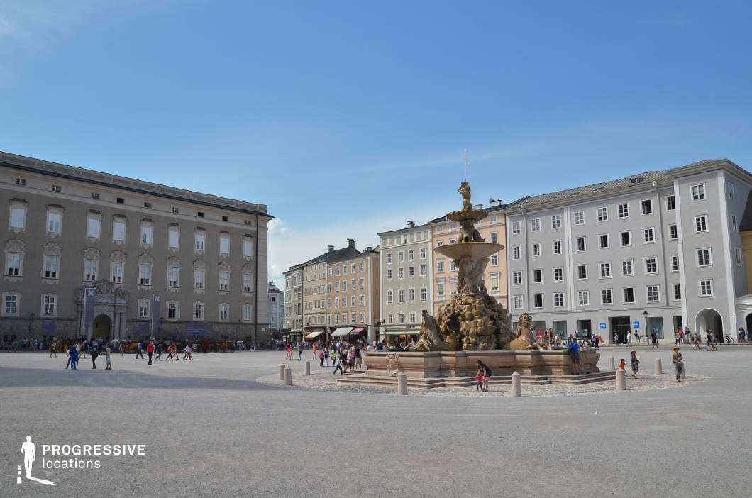 This spacious urban square is dominated by a monumental baroque fountain featuring dynamic sculptural figures and flowing water, set at the center of an open gravel plaza. Surrounding the square are uniform façades of multi-story buildings in pastel and neutral tones, with simple geometric windows and minimal ornamentation that create a clean and orderly backdrop. The architecture spans classical and early modern styles, with subtle decorative reliefs enhancing the historic atmosphere. The composition emphasizes symmetry and grandeur, with broad sightlines and an open layout that enhances depth and movement. This location is ideal for historical or period dramas, cultural documentaries, or commercial shoots requiring a stately, European urban environment.