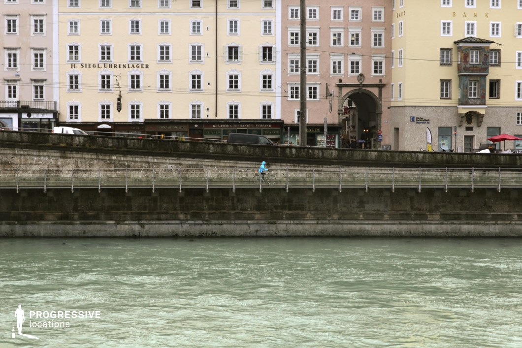 This image presents a clear view of a European urban riverside, with a tiered embankment built from aged stone and topped by a metal-railed pedestrian and cycling path. Behind it, multi-story Baroque and classical-style façades in pastel tones—light yellows, creams, and muted pinks—rise in a grid of regularly spaced windows. A prominent arched gateway and a painted oriel window add architectural depth and focal interest. The composition features strong horizontal layering: the river, the retaining wall, the walkway, and the buildings, all stacked with clarity. The perspective from across the water offers a balanced, symmetrical framing, emphasizing the location’s architectural regularity and historical texture. This setting is ideal for urban scenes requiring a distinctly European waterfront, suitable for genres like drama, art films, or travel and lifestyle productions focusing on classical city environments.