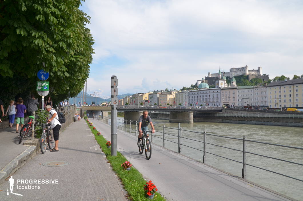 This urban riverside promenade features a dual-level path system with separate lanes for pedestrians and cyclists, bordered by neat rows of flower planters and a sturdy metal railing along the water's edge. To the left, mature tree canopies provide natural shading over a paved walkway, while on the opposite bank, a cohesive row of pastel-colored historic buildings lines the riverfront. Dominating the skyline is a hilltop fortress with white stone walls and towers, offering a strong vertical contrast and historical depth. The bridge connecting the two sides enhances the spatial layering and urban continuity. The image is captured from a street-level angle, creating a linear visual flow that draws the eye along the river and into the cityscape. This location is ideal for filming contemporary urban scenes, establishing shots, or travel-related content, and it suits genres such as drama, romance, or light-hearted city explorations.