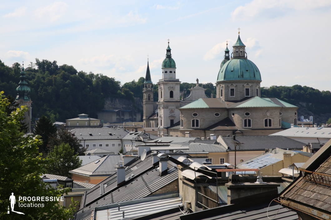 This elevated cityscape reveals a tightly packed array of pitched rooftops in muted greys and browns, contrasted by a backdrop of prominent Baroque architecture, including a large domed cathedral with green patina copper roofing and several ornate church towers. The layered rooflines create strong diagonal geometries that draw the eye upward toward the skyline, where the historic towers rise above the urban fabric. Architectural details such as clock faces, domed spires, and decorative lantern towers enhance the classical European atmosphere. A wooded hillside in the background adds natural depth, while the mid-height perspective balances foreground texture with architectural grandeur. This location is ideal for filming scenes that require an old European town ambiance, and it suits genres such as historical drama, period romance, or travel and culture-focused content.