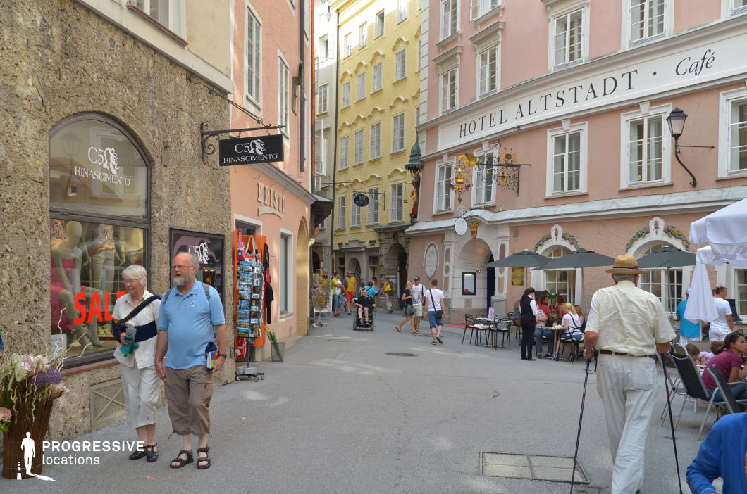 This lively pedestrian alley is set within a historic urban core, featuring narrow walkways lined with pastel-hued facades in shades of pink, peach, yellow, and beige. The buildings are mid-rise and stylistically consistent, with traditional window arrangements and arched ground-floor entrances. Boutique signage, wrought-iron mounts, and hanging decor contribute to the authentic European old-town charm. One corner hosts an outdoor café with neatly arranged seating under dark umbrellas, creating a welcoming and inhabited atmosphere. The street surface is smooth and neutral, allowing architectural and retail details to stand out. With its visually layered facades, tight scale, and human-focused design, this location is ideal for commercial shoots, travel segments, or films needing a cozy, culturally rich urban backdrop.