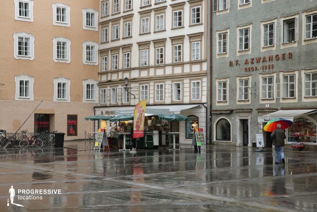 This image depicts a small urban square bordered by mid-rise historic buildings featuring varied pastel façades, ornate window frames, and traditional European plaster textures. The square’s surface is composed of large, reflective stone slabs that create clean visual lines and mirror the architectural details, enhancing depth and symmetry. The buildings range from soft peach and ivory tones to muted greens, presenting a visually rich yet cohesive palette. Ground-level commercial kiosks with awnings introduce an informal, functional layer to the space, while the upper stories maintain a classical, structured rhythm. The compact scale of the square, combined with the intricate architectural elements, provides an intimate and textured urban backdrop. This setting is especially suited for character-driven dramas, romantic scenes, or European city montages requiring a cozy, historically rich environment with strong vertical and planar visual elements.
