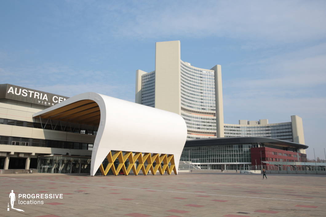This architectural complex features a combination of modernist and contemporary design elements, with a striking curved white canopy supported by a geometric yellow framework at the forefront. The large, open plaza is composed of a patterned tiled surface, adding depth to the setting. In the background, a towering high-rise with a curved glass facade reflects the surrounding environment, reinforcing the location’s corporate and institutional aesthetic. The mix of curved and angular structures creates a visually dynamic composition, making this an ideal filming location for corporate, political, or espionage-themed productions. The expansive open space is well-suited for establishing shots, public addresses, or high-stakes negotiation scenes.