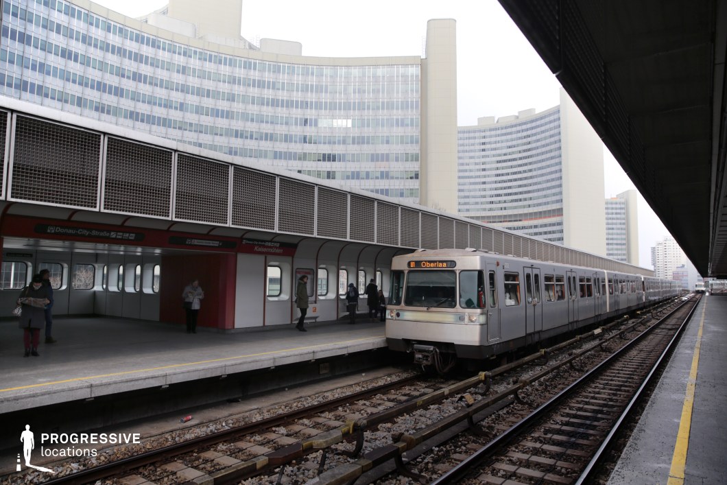 This metro station presents a blend of modern infrastructure and functional design, with a long, elevated platform covered by metal paneling and industrial-style ventilation grids. The station's red and white color scheme contrasts with the silver metro train, which has a streamlined, utilitarian appearance. In the background, a curved glass-and-concrete high-rise complex dominates the skyline, adding a sleek, corporate aesthetic to the urban environment. The station’s symmetrical layout and converging railway tracks create a strong sense of depth and perspective. This setting is ideal for filming scenes in genres like sci-fi, thrillers, or espionage films, where public transportation hubs serve as key locations for dramatic arrivals, tense departures, or surveillance sequences.