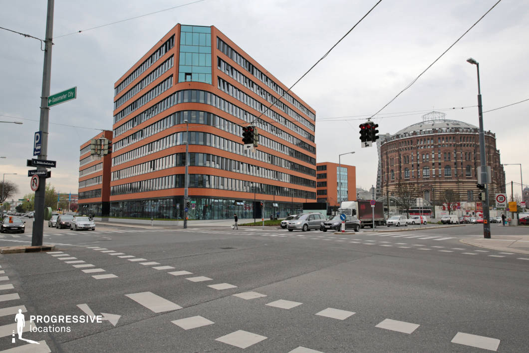 This urban setting showcases a striking contrast between contemporary and historic architecture. The foreground features a wide intersection with clearly marked pedestrian crossings, traffic signals, and overhead tram cables, indicating a well-connected transportation hub. The modern glass and brick office building has a dynamic, angular design with curved elements, blending warm brick tones with reflective windows. In the background, a cylindrical historic structure with an industrial aesthetic, possibly a former gasometer, stands out with its ornate brickwork and metal detailing. The scene’s mix of architectural styles and its functional urban layout make it ideal for filming corporate, crime, or dystopian-themed sequences, as well as establishing shots for a bustling city environment.