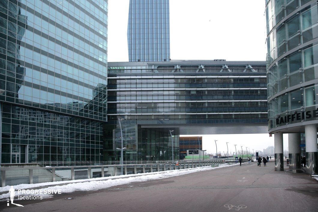 This modern cityscape features towering glass and steel structures, reflecting the surrounding environment in their sleek, mirrored facades. The geometric arrangement of buildings, with a suspended bridge-like structure connecting two sections, creates a futuristic and corporate aesthetic. The clean lines and metallic surfaces contrast with the open pedestrian space below, which is flanked by contemporary streetlights and minimalistic urban infrastructure. This location is ideal for corporate dramas, high-tech thrillers, or dystopian science fiction, offering a polished, futuristic setting that evokes themes of power, innovation, and modernity.