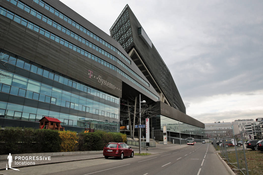 This imposing modern office complex features a striking angular design with dark, reflective glass and metal panels that create a sleek, futuristic aesthetic. The bold architectural lines, supported by diagonal beams and open structural elements, enhance the building’s dynamic and avant-garde character. A wide, gently sloping street runs alongside, flanked by a mix of commercial and office buildings, adding to the contemporary urban setting. The combination of sharp geometric forms and expansive glass surfaces makes this location ideal for filming corporate thrillers, dystopian sci-fi, or high-stakes business dramas, conveying a sense of power, innovation, and modernity.
