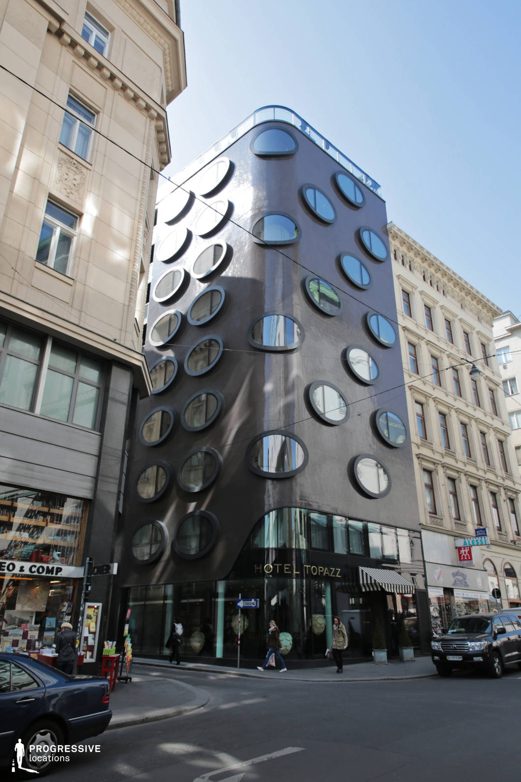 This striking modern building stands out with its dark, glossy façade punctuated by a grid of large, round windows, creating a futuristic yet organic aesthetic. The curved edges and reflective surfaces contrast sharply with the surrounding traditional European architecture, emphasizing its bold, avant-garde design. The ground level features a sleek glass storefront with elegant signage, integrating seamlessly into the urban streetscape. The juxtaposition of old and new architectural styles makes this location ideal for filming contemporary dramas, fashion editorials, or high-concept sci-fi and mystery productions, offering a visually distinctive and unconventional city setting.