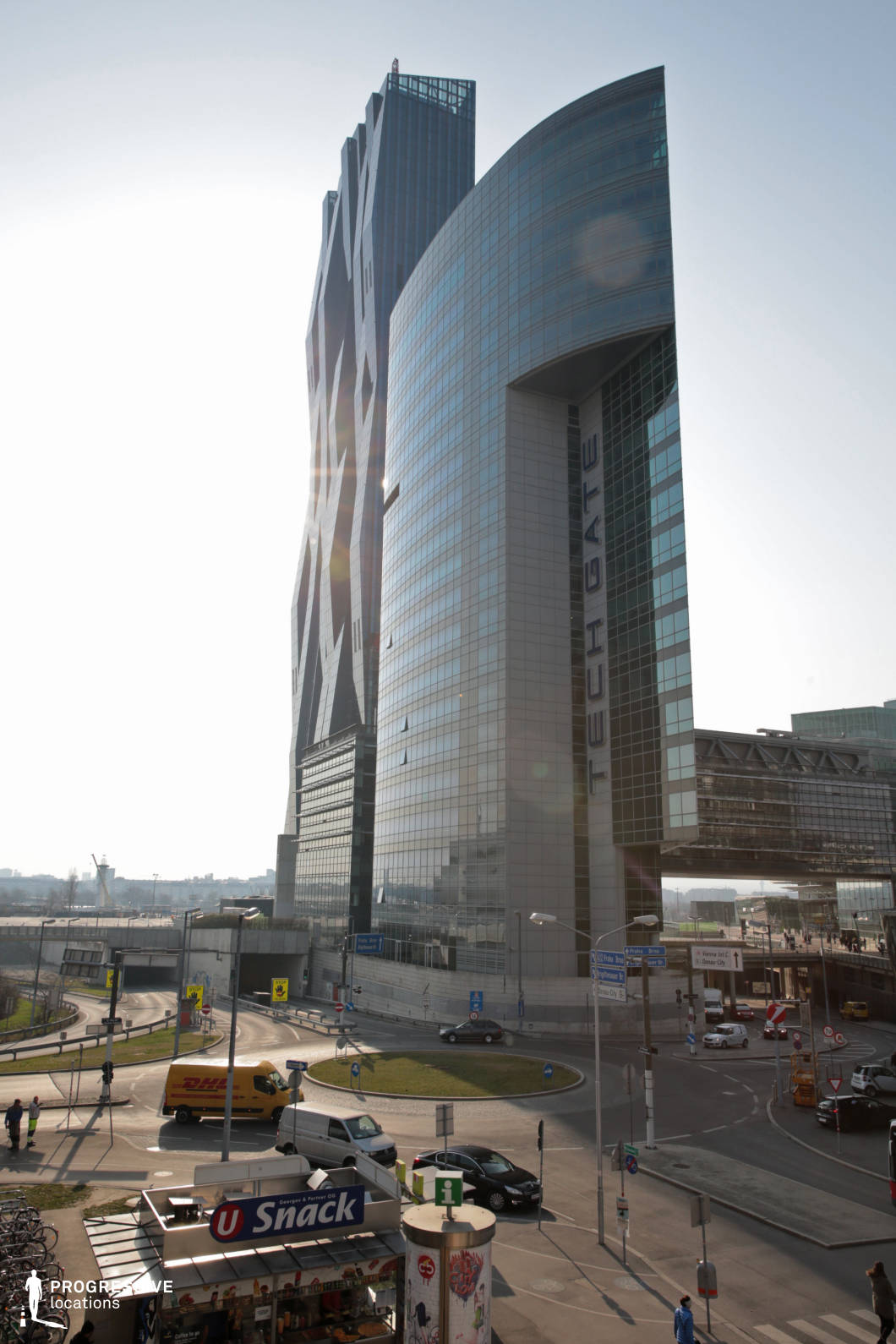 This modern business district showcases a striking ensemble of contemporary high-rises with bold architectural designs. The dominant structures feature sleek glass facades reflecting the surrounding cityscape, with one tower boasting a dynamic, angular pattern that adds a futuristic aesthetic. The curved and linear elements of the buildings create a visually compelling contrast, further enhanced by an elevated pedestrian bridge connecting different sections of the complex. The surrounding infrastructure includes multilane roads, roundabouts, and urban signage, emphasizing the area’s functionality and fast-paced nature. This location is ideal for corporate thrillers, cyberpunk-inspired sci-fi, or high-stakes business dramas, offering a sophisticated and forward-thinking setting for dynamic storytelling.
