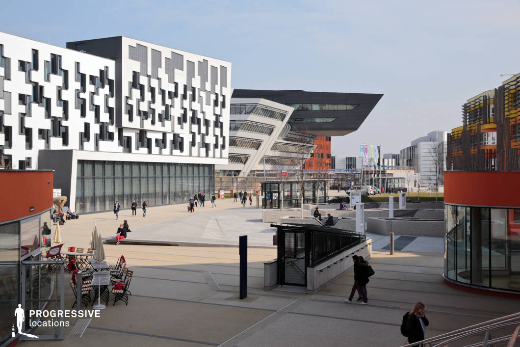 This modern campus blends avant-garde architectural styles, featuring angular and geometric facades, cantilevered structures, and vibrant color contrasts. The striking black overhanging building in the background, with its sharp lines and expansive glass surfaces, dominates the skyline, while the pixelated facade of the adjacent structure adds a sense of dynamism. The open plaza is designed with wide pedestrian pathways, sunken seating areas, and integrated landscaping, creating a contemporary and inviting urban space. The mix of materials, from smooth concrete to metal and glass, enhances the futuristic aesthetic. This location is ideal for sci-fi films, high-tech thrillers, or academic dramas, providing a sleek and visionary backdrop for intellectual discourse, futuristic scenarios, or corporate intrigue.