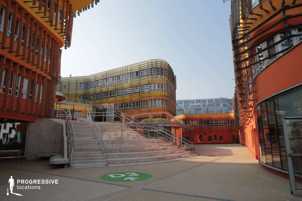 This location showcases a striking contemporary architectural design, featuring curved and layered facades adorned with wooden slats and metal frameworks. The buildings incorporate warm hues of orange and yellow, creating a dynamic contrast against the neutral tones of the concrete stairways and walkways. The spacious courtyard is framed by curved glass windows and interconnected staircases, enhancing depth and movement within the space. The setting is ideal for filming scenes that require a futuristic, academic, or high-tech corporate environment. Its blend of organic and industrial elements makes it suitable for genres like sci-fi, drama, or psychological thrillers, with potential use for establishing shots, corporate intrigue, or university-based narratives.